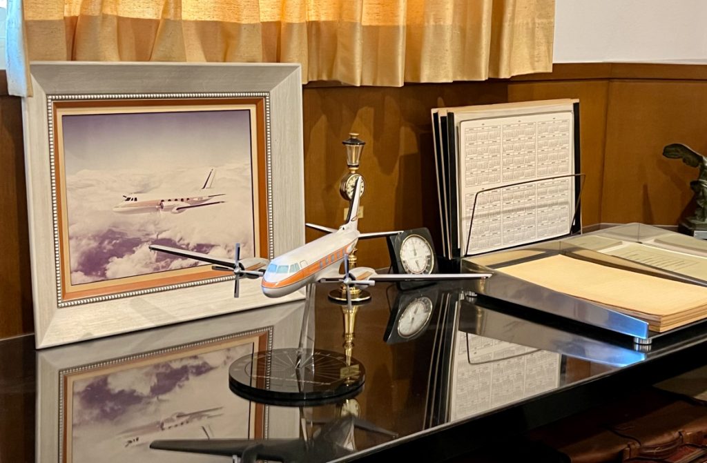 A small model of Walt’s Plane is mounted on a stand. The plane is white with an orange stripe and blue windows. Behind the model plane is a photo of the plane in the sky bordered by a white frame. The plane and photo are sitting on top of a glass-topped desk. On the desk are other items such as a small clock and documents.