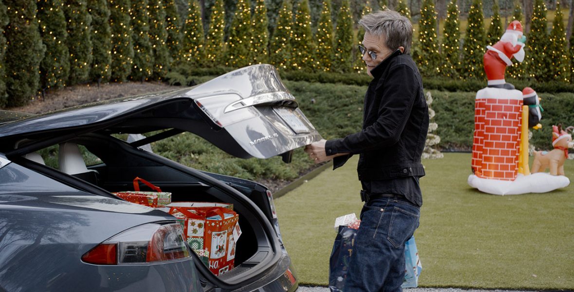 Kevin Bacon lifts the trunk of his car, revealing multiple Christmas presents. He is wearing a black jacket, blue jeans, and sunglasses, and he holds two bags in his hands. Behind him is a nicely manicured lawn and dozens of lit-up Christmas trees. An inflatable Santa Claus coming down the chimney—along with a penguin and a reindeer—is on display in the yard.