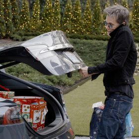 Kevin Bacon lifts the trunk of his car, revealing multiple Christmas presents. He is wearing a black jacket, blue jeans, and sunglasses, and he holds two bags in his hands. Behind him is a nicely manicured lawn and dozens of lit-up Christmas trees. An inflatable Santa Claus coming down the chimney—along with a penguin and a reindeer—is on display in the yard.
