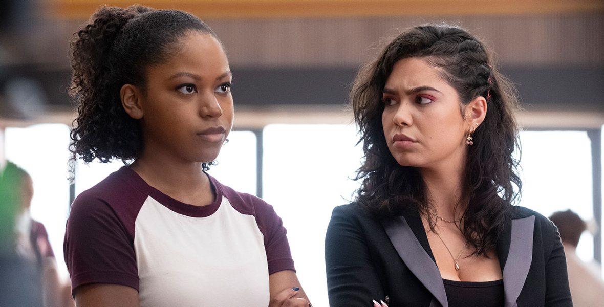 Darby (Riele Downs) wears a white and maroon t-shirt. She has curly hair that is tied behind her head. Capri (Auli’i Cravalho) is on her right, looking at her with her arms crossed. Capri is wearing a black and gray blazer. She has wavy hair that is tucked behind her left ear.