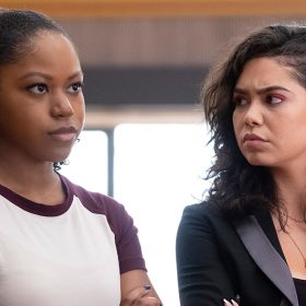 Darby (Riele Downs) wears a white and maroon t-shirt. She has curly hair that is tied behind her head. Capri (Auli’i Cravalho) is on her right, looking at her with her arms crossed. Capri is wearing a black and gray blazer. She has wavy hair that is tucked behind her left ear.