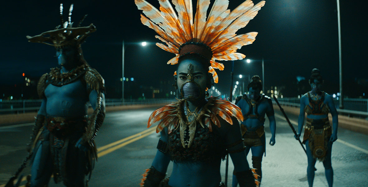 Namora (Mabel Cadena), a blue-skinned woman wearing a breathing mask and a collar and headdress inspired by a lionfish, walks along a street at nighttime. Behind her are other blue-skinned warriors, following her as they walk towards the camera.