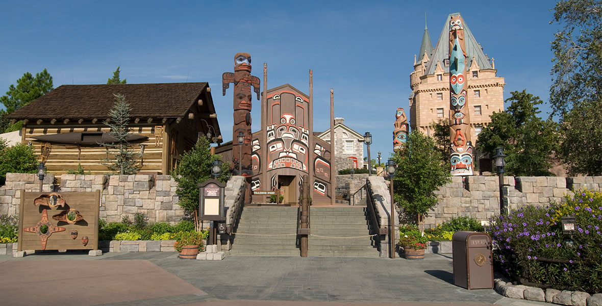A wide shot of the Canada Pavilion at EPCOT. It is sunny outside and the sky is clear blue. A wooden cabin, large caved totem poles, and a castle-like building are visible.