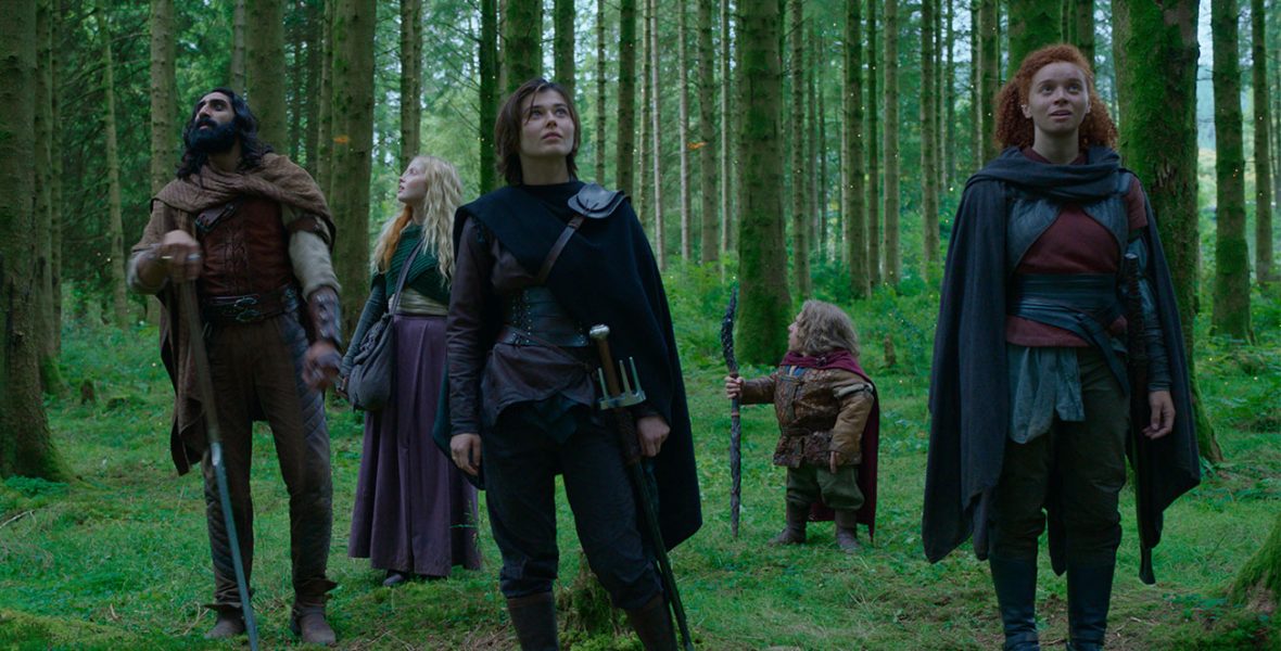 In a scene from the Disney+ Original series Willow, the stars of the series stand in a lush, green forest with trees covered in moss and ivy. From left to right are actors Tony Revolori as Graydon, Amar Chadha-Patel as Boorman, Ellie Bamber as Dove, Ruby Cruz as Kit, Warwick Davis as Willow Ufgood, and Erin Kellyman as Jade. Revolori wears black knee-high boots, black pants, a black long-sleeved top, and a charcoal hooded vest. Chadha-Patel wears brown boots, brown pants, a green hooded long-sleeved top, and holds a large metal sword in his right hand. Bamber wears a purple skirt and green knitted sweater. Cruz wears a dark long-sleeved top, black pants, black knee-high boots, and a leather corset. Davis holds a wooden staff in his right hand and wears a maroon cape, olive pants, and brown leather backpack. Kellyman wears olive pants, black knee-high boots, a gray long-sleeved top with a maroon short-sleeved top, and a leather vest.