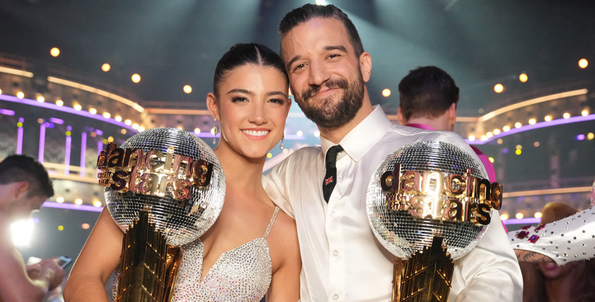 Charli D’Amelio (left) and Mark Ballas (right) smile for a photo while holding their Mirrorball trophies. D’Amelio wears a bedazzled silver and yellow dress, with her hair pulled back into a tight bun. Ballas wears bedazzled pinstripe pants, a white dress shirt, and a black tie with a British flag pin.