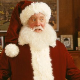 Tim Allen is dressed in Santa Claus’ classic red velvet suit with white fur trim and a matching cap. He has a long white beard and has his hands out on either side of him.