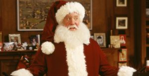 Tim Allen is dressed in Santa Claus’ classic red velvet suit with white fur trim and a matching cap. He has a long white beard and has his hands out on either side of him.