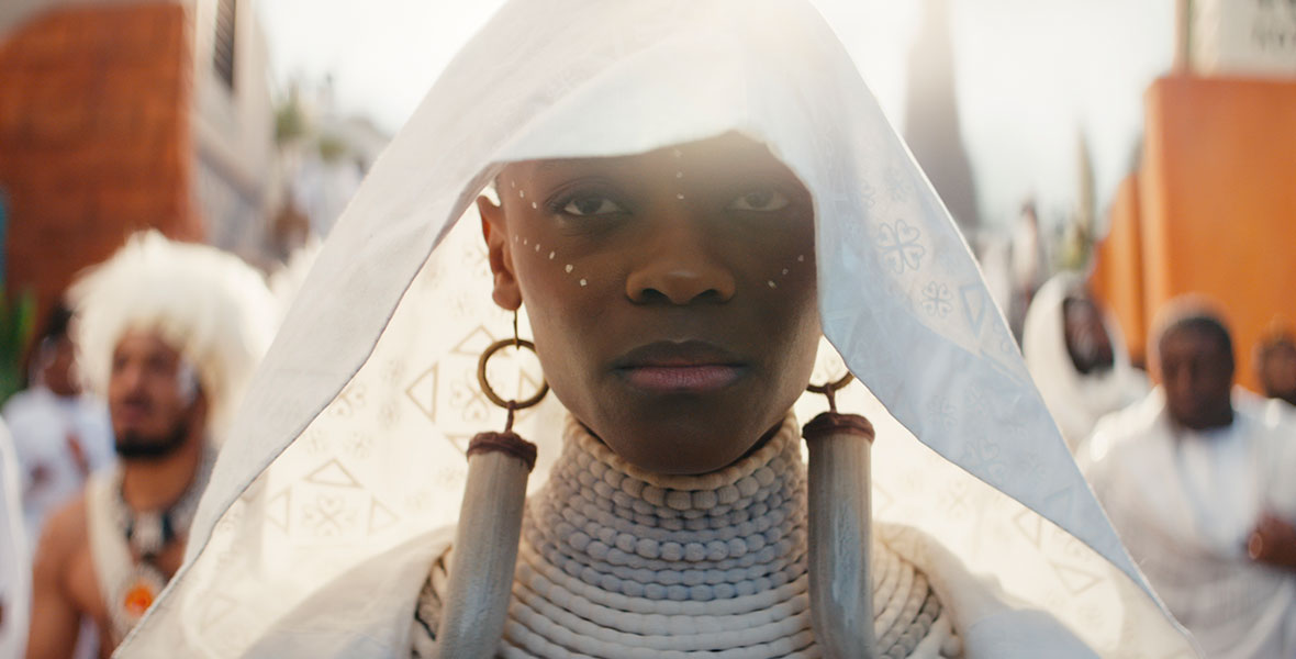 In a still from Black Panther: Wakanda Forever, Shuri (Letitia Wright) wears white ceremonial funeral garb and makeup. She has a sad expression on her face. In the background are mourners, also wearing white.