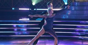 Selma Blair and pro Sasha Farber perform a contemporary routine. A barefoot Blair (front) wears a bedazzled blue leotard with cut-out details. Farber (rear) holds Blair’s hands, which are fully extended on each side.