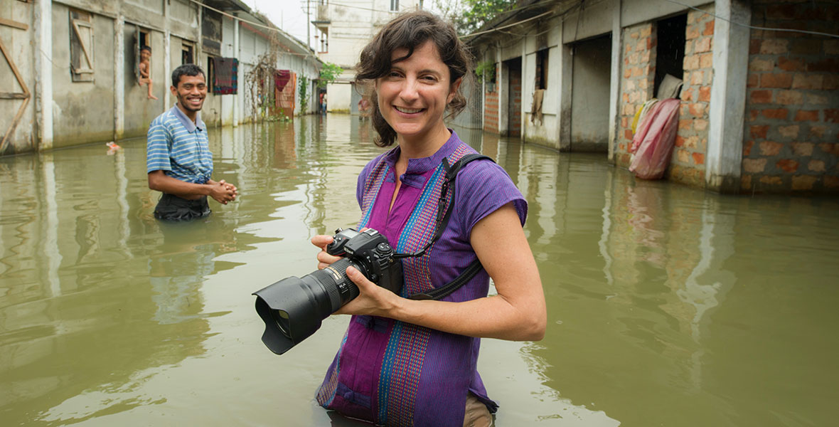 Ami Vitale stands in an alley between buildings holding her camera. She is submerged in green murky water that reaches all the way up to her hips and she is wearing a purple patterned shirt and smiling at the camera. Behind her is a man wearing a blue striped polo shirt, also smiling, and behind him there is a blurry image of a child hanging out of a building window. 