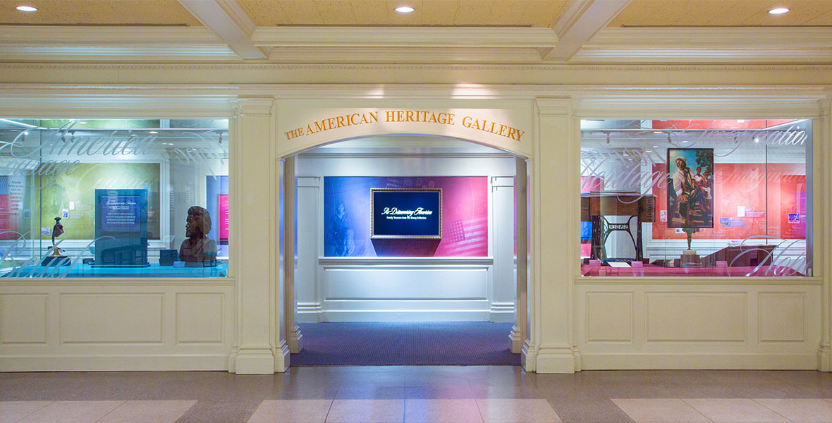 Wide shot of The American Heritage Gallery at EPCOT. There are various art pieces inside glass display cases on either side of a doorway with “The American Heritage Gallery” above the door in gold lettering. Inside the exhibit are red, white, and blue murals and screens.