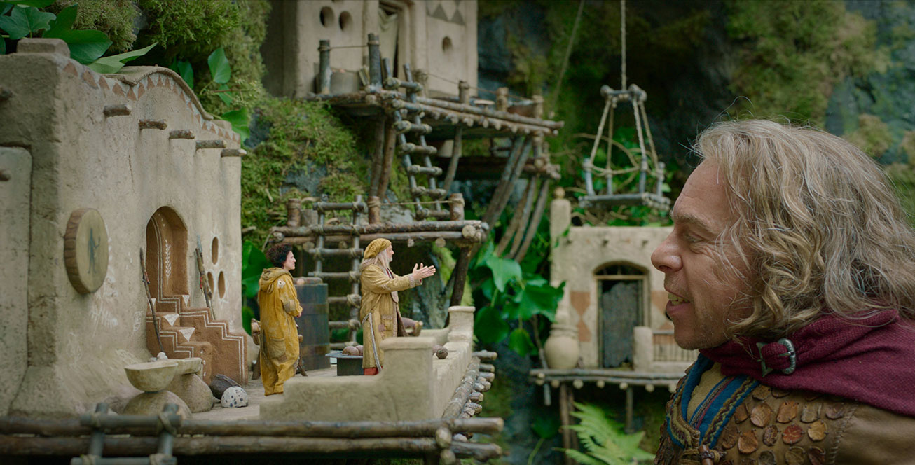 In a scene from the Disney+ Original series Willow, Warwick Davis portrays Willow Ufgood. He wears a brown top with a maroon cape draped around his neck. He smiles and stands next to the home of the Brownies—who wear tan tunics and stand on a small balcony. Small mud structures are adorned with wooden ladders and sit in a large tree.