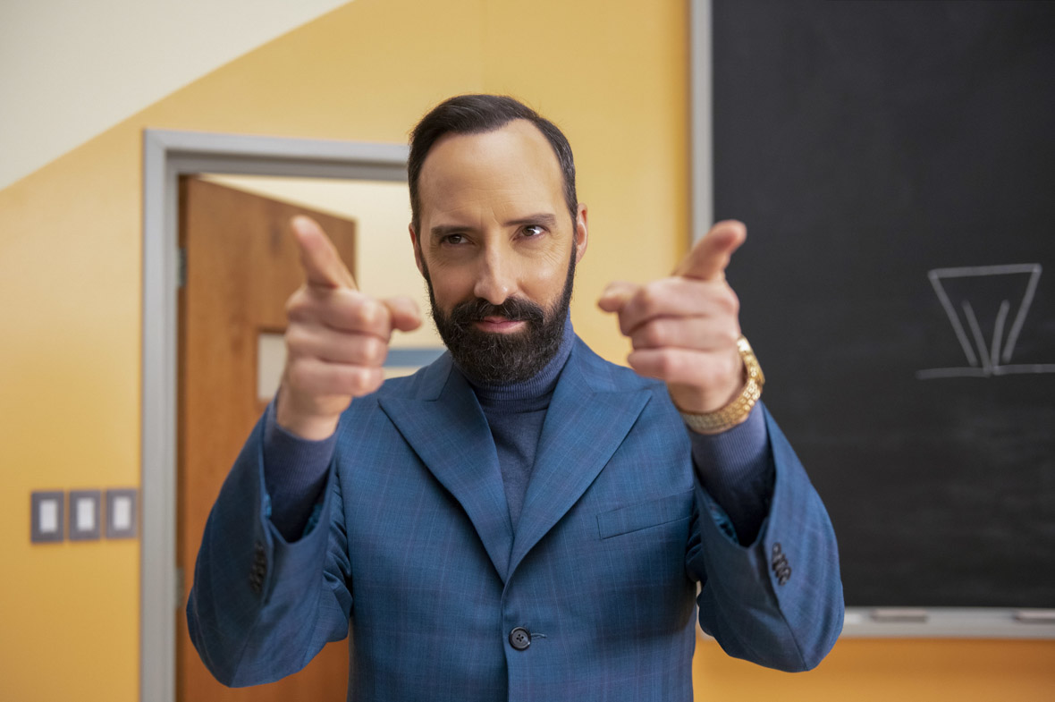 Mr. Benedict (Tony Hale) wears a blue suit in a scene from The Mysterious Benedict Society. He is bearded and smirking, and he is raising his arms, extending his pointer fingers. He is standing in a classroom that has yellow walls and a black chalkboard.
