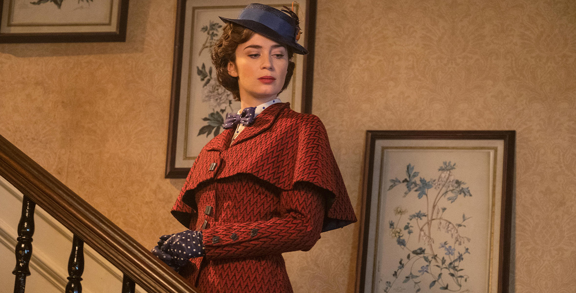 In a scene in Mary Poppins Returns, actor Emily Blunt portrays Mary Poppins. She wears a matching red suit jacket and skirt, a navy bowtie with white polka dots, a white blouse with navy polka dots, and navy gloves with white polka dots. She stands on a staircase and looks to the foot of the stairs. Three paintings of flowers hang on the wall behind her in brown wooden frame.
