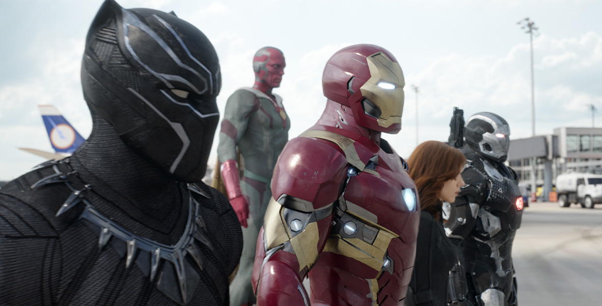 From left to right, Black Panther, Vision, Iron Man, Black Widow, and War Machine stand side-by-side. The tail of a plane is seen behind them with a blue and white logo. A large white truck is on the far right. Actor and Disney Legend Chadwick Boseman portrays Black Panther and wears an all-black suit with his face covered. Actor Paul Bettany portrays Vision with a charcoal suit and red face. Actor and Disney Legend Robert Downey Jr. portrays Iron Man and wears a red metal suit with gold accents. Actor Scarlett Johansson portrays Black Widow and wears an all-black suit. Actor Don Cheadle portrays War Machine and wears a black and silver metal suit.