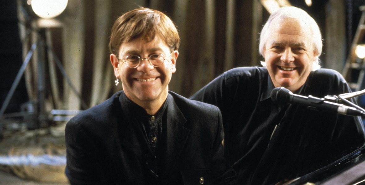 Musicians and Disney Legends Elton John and Tim Rice sit on a bench next to a black piano. John wears an all-black suit with a black button-down dress shirt and thin, wire-framed glasses. Rice leans on the piano and wears a black sweater and dark charcoal button-down shirt. A black microphone is propped on the piano and rests near Rice’s mouth. Behind them are long, tan curtains and large lighting fixtures.