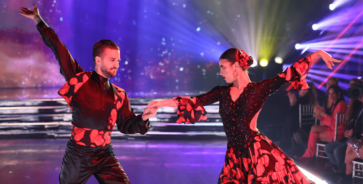 In a scene from an episode of the Disney+ series Dancing with the Stars, TikTok star Charli D’Amelio and partner pro Mark Ballas perform a dance routine. Ballas wears a black button-down shirt and black pants with a red patterned vest and cummerbund. D’Amelio wears a black and red gown that matches Ballas’ vest and cummerbund. Ballas and D’Amelio stand with their outside arms extended upward. Bright spotlights shine on Ballas and D’Amelio and a screen in the background displays a galactic scene.