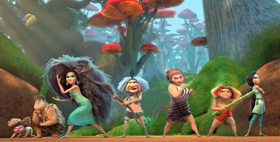 In a scene from the series The Croods: Family Tree, Gran stands center and holds a long wooden stick. She wears a bikini made from turtle shells and bones, with green paint on her face. On each side stands other female members of the Crood family with colorful paint on their faces and prehistoric outfits. Each stands ready to attack an enemy with their hands in fists. Behind them is a dense, colorful forest with abstract-shaped plants and a bright blue sky.