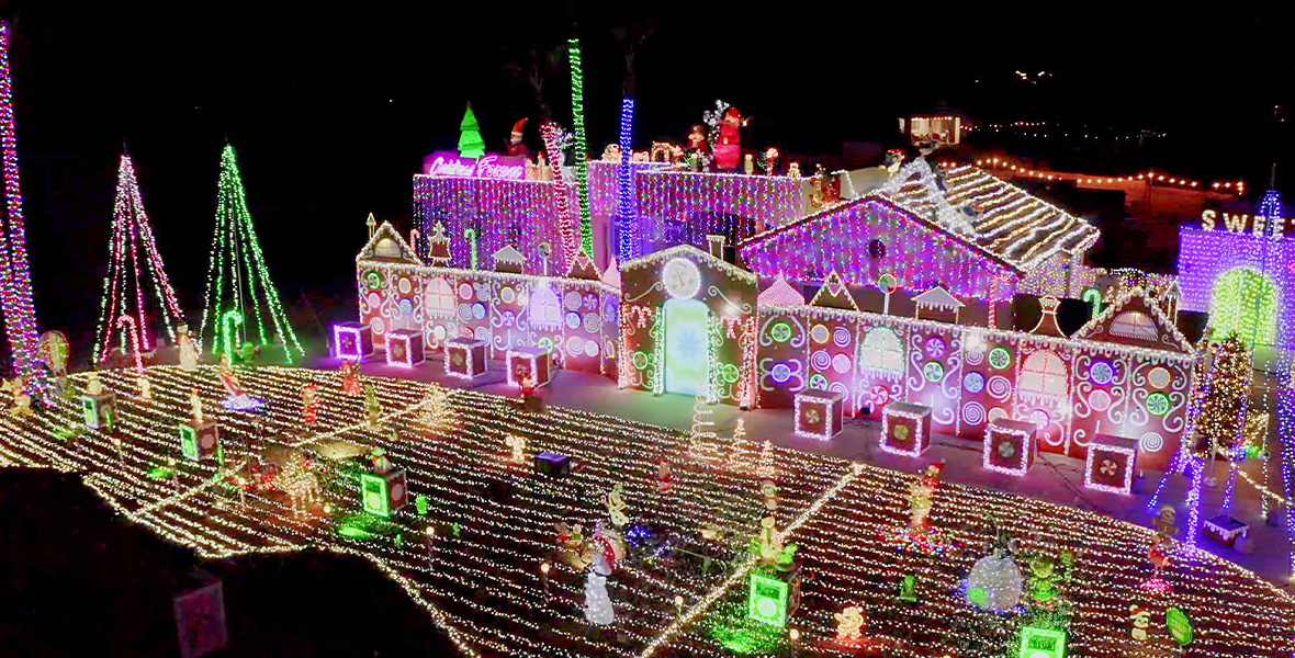 In a scene from The Great Christmas Light Fight, an aerial shot shows a residential house adorned with thousands of white and colored holiday lights. The front yard is covered with light strands and inflatable decorations of snowmen, Santa Claus, Olaf from Disney’s Frozen, and more. On the side of the house are large trees made of light strands. On the roof are lighted decorations including Christmas trees, Santa Claus, candy canes, and more.