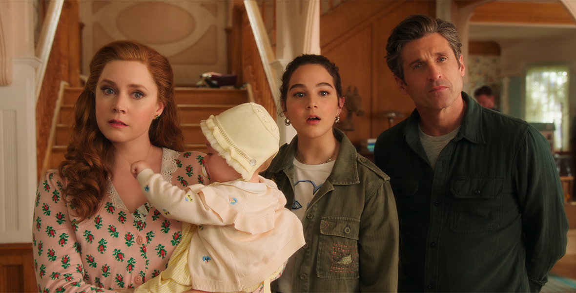 In a production still from Disenchanted, Giselle (Amy Adams), Morgan (Gabriella Baldacchino), and Robert (Disney Legend Patrick Dempsey) are standing in their new house; they have quizzical looks on their faces, and are looking just off camera. Giselle is holding baby Sophia. A stairway is seen behind them, as are a few workmen to their right.
