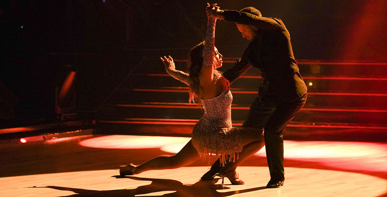 Cheryl Burke (left) wears a sequined, fringed dress with matching gloves. Her right knee is bent and her left leg is fully extended behind her. Both of Burke’s arms are in the air, and one hand is clasping Pasha Pashkov’s hand. He is slightly bent over, facing Burke. They are cast in a red light on the ballroom floor.