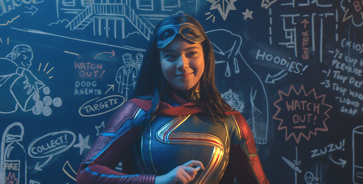 Kamala Khan is wearing her Ms. Marvel suit, which is red, blue and gold. Behind her is a chalk board with several illustrations. She is holding a piece of chalk.