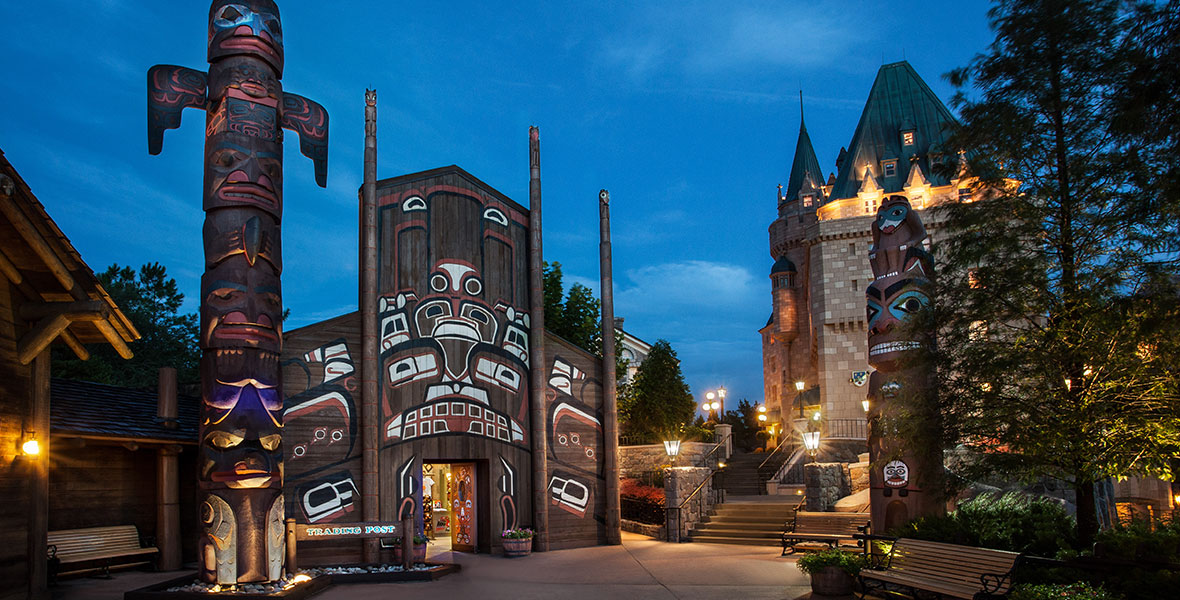 A nighttime shot of the trading post at EPCOT’s Canada Pavilion. The hand carved totem poles are illuminated with up-lighting and the door to the Trading Post store is open with light shining from inside.