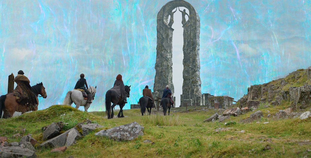 In a scene from the Disney+ Original series Willow, the stars ride horses on grassy terrain with scattered boulders, toward a large stone archway with two human sculptures decorating the top.