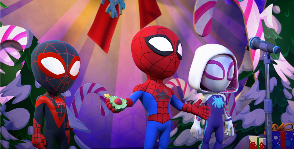 In a scene from the animated series Marvel’s Spidey and his Amazing Friends, Spider-Man/Spin/Miles Morales, Spider-Man/Peter Parker, and Ghost-Spider/Gwen Stacy stand side-by-side on a stage and behind a microphone. Peter stands with his arms extended and holds a Christmas tree-shaped object in his right hand. The stage is decorated with large cutouts of candy canes, Christmas trees, and snowflakes, and large presents are placed on the floor to the left of Gwen.