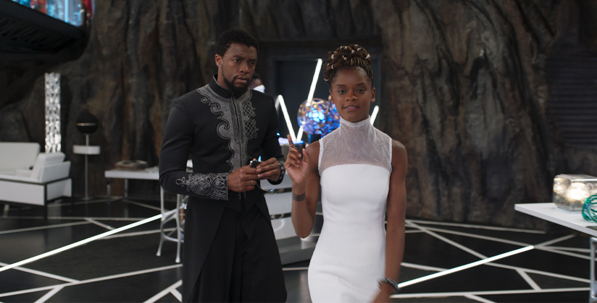 Actor and Disney Legend Chadwick Boseman portrays T’Challa/Black Panther. Boseman wears a long-sleeved traditional African embroidered robe and holds a small remote in his hands. Actor Letitia Wright portrays Shuri and wears a white dress with a high mesh neckline. Boseman and Wright stand on a black floor with geometric designs. A white couch and chair are to their right while a long white table with knickknacks is on the left.