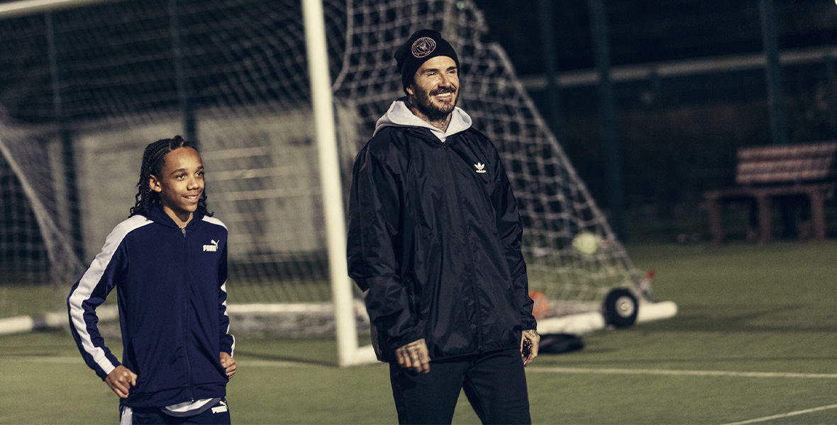 In a still from Disney+’s Save Our Squad with David Beckham, from left to right, a young boy stands with his hands on his hips. He wears a navy-blue zippered jacket with a thick white stripe down the arms and matching pants. Former soccer star David Beckham stands to the right and smiles; he wears a black beanie with a large logo, black windbreaker jacket, black pants, and a gray hoodie. They stand on a soccer field with a large goal behind them.