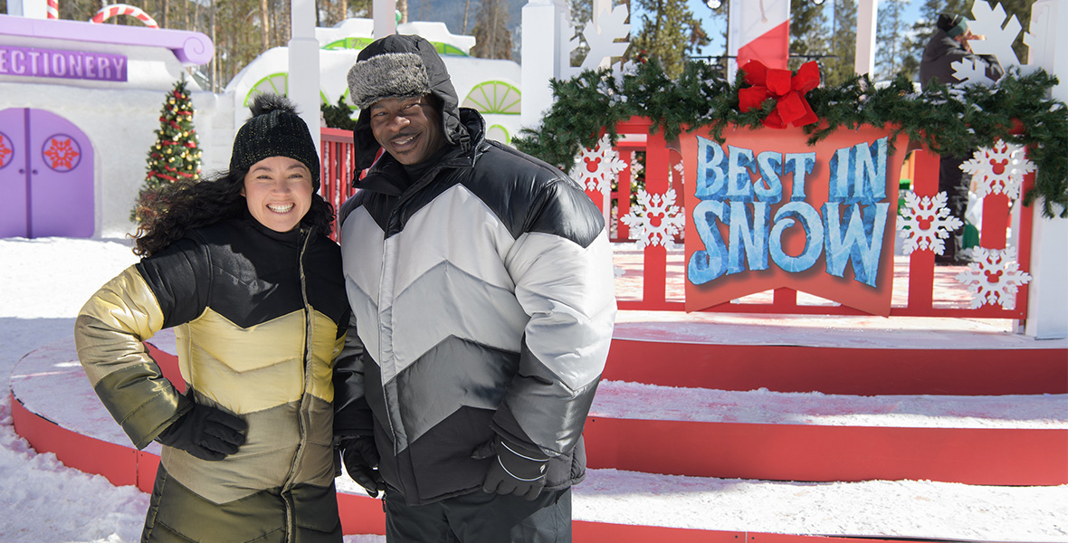 From the special Best in Snow, judges Sue McGrew and Andre Rush pose side by side. Behind them is a large stage with red steps and covered in snow. A sign on the stage reads, “BEST IN SNOW” in blue writing on a red background. Christmas trees and garlands decorate the area behind them.