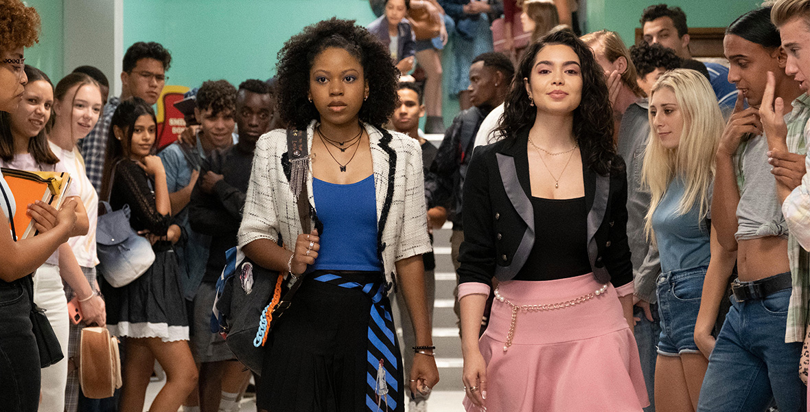 In a scene from 20th Century Studios’ Darby and the Dead, actors Riele Downs as Darby Harper and Auli’i Cravalho as Capri walk side by side down a high school hallway. Downs wears a cream blazer, a blue tank top, a dark blue skirt, black knee-high socks, and a blue and black striped tie tied around her waist. Cravalho wears a black blazer with satin lapels, a black tank top, and a pink skirt. Downs and Cravalho are surrounded by a crowd of high school-aged students who are all staring at them.