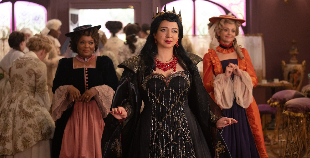In a production still from Disenchanted, Rosaleen (Yvette Nicole Brown), Malvina (Maya Rudolph), and Ruby (Jayma Mays) are standing and looking off camera. There is a group of people clumped together behind them, exiting through a door. Rosaleen (on the left) is wearing a black and pink gown with a black hat; Malvina (in the middle) is wearing a black gown with gold beaded trim and a black cape, as well as a red beaded necklace and a gold crown; and Ruby (on the right) is wearing an orange and purple gown with an orange hat. The three have smirks on their faces.