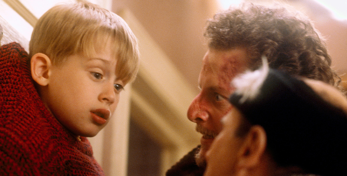 A young boy, Kevin McCallister is pinned up against a wall by two thieves. He is wearing a red knitted sweater and has blonde hair. The thieves are staring at him.