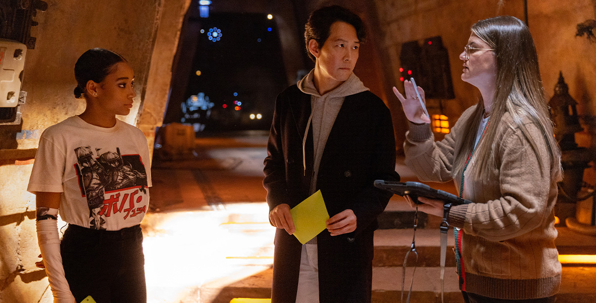 A behind-the-scenes image from production of Lucasfilm’s The Acolyte, coming to Disney+. Creator Leslye Headland, on the right, is holding a tablet computer and gesturing; she’s wearing a brown sweater and has long brown hair. In the middle is actor Lee Jung-jae; he’s wearing a black coat with a gray hooded sweatshirt underneath. On the left is Amandla Stenberg; she’s wearing a white t-shirt with a graphic on it and black pants. Stenberg and Lee are looking at Headland.