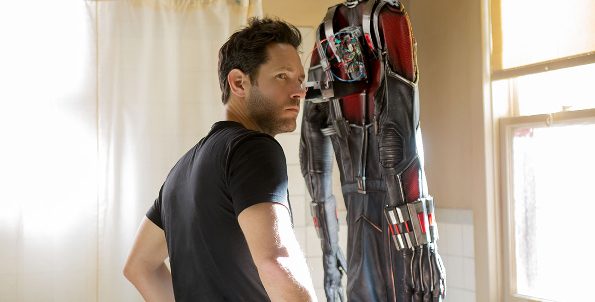Scott Lang wears a black T-shirt looking to over his right shoulder. His Ant-Man suit is hanging on a shower curtain rod in front of him.