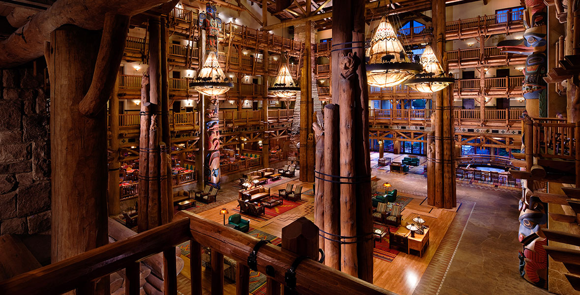 Wide view of the Wilderness Lodge lobby. Large, themed chandeliers hang from the ceiling with silhouettes of bison and other animals. Large wooden poles hold up the ceiling and carved totem poles adorn the walls. The lobby is empty, but you can see chairs, couches, and rugs available for guests to sit on.