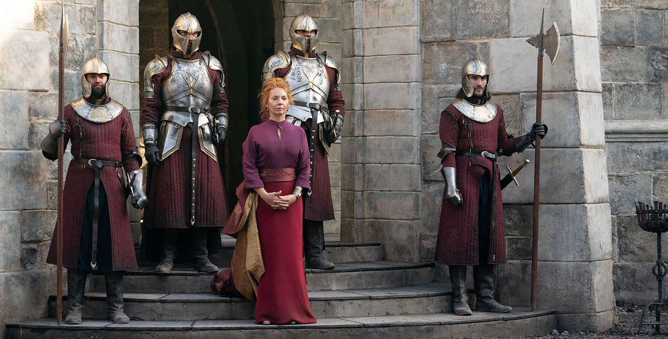 In a scene from the Disney+ Original series Willow, actor Joanne Whalley portrays Sorsha; she stands on stone steps and is surrounded by four men wearing metal armor and maroon tunics. She wears a purple blouse and a cardinal maxi skirt with maroon and gold fabric wrapping her waist and draping down her side. Behind them is a tall stone archway; in front of them, a gravel pathway.