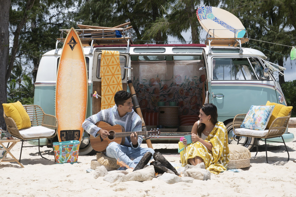In this scene from Doogie Kamealoha, M.D., Walter (Alex Aiono) strums a guitar while sitting on the beach. Behind him is an orange surfboard, a blue camper van, a wicker chair and table set, and a tote bag. Next to him is Doogie (Peyton Elizabeth Lee), who is wrapped in yellow towel and holding a blue mug. In front of both is a fire pit.