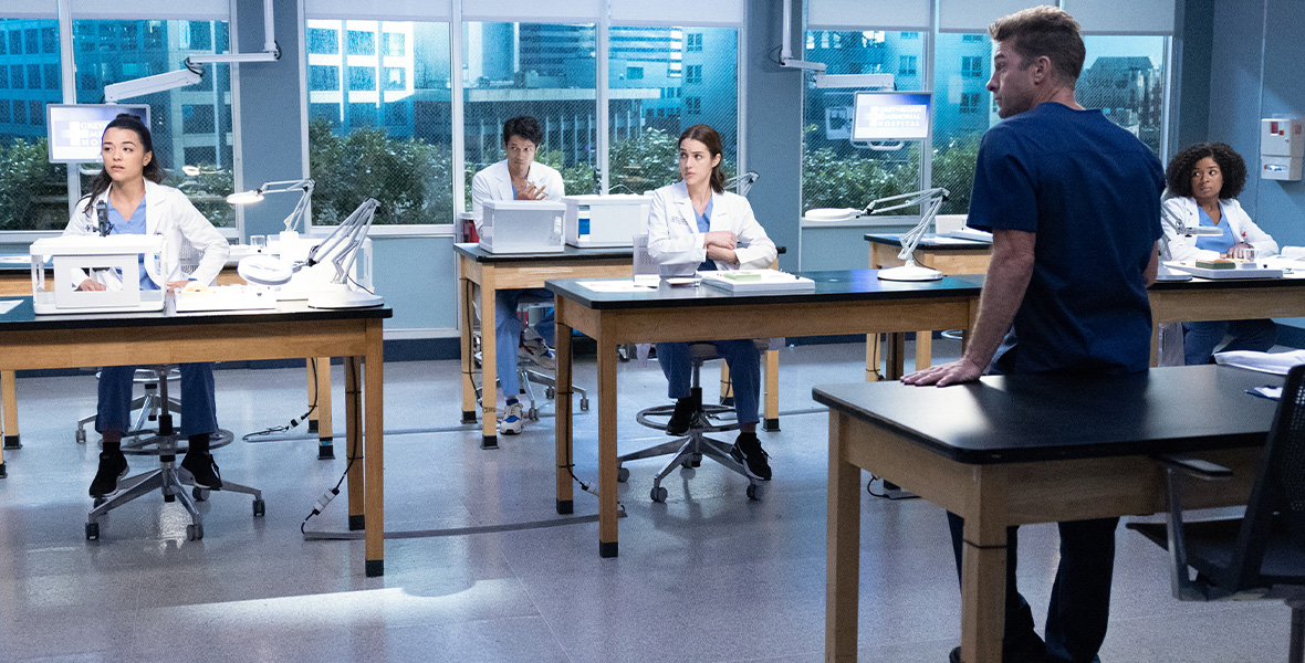 In a still from ABC’s Grey’s Anatomy, from left to right, actors Midori Francis, Harry Shum Jr., Adelaide Kane, and Alexis Floyd each sit solo at large desks. Francis, Shum, Kane, and Floyd wear white medical coats and blue scrubs. The tables are covered with large white machinery and metal lights. Actor Scott Speedman stands facing the group and leans on a desk. He wears dark blue scrubs.