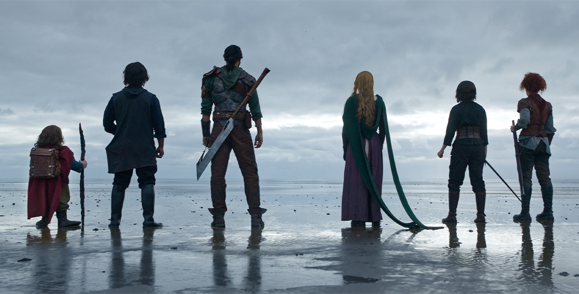 In a scene from Disney+ Original series Willow, the stars stand with their backs toward the viewers as they stare at a large body of water. From left to right stand actors Warwick Davis as Willow Ufgood, Tony Revolori as Graydon, Amar Chadha-Patel as Boorman, Ellie Bamber as Dove, Ruby Cruz as Kit, and Erin Kellyman as Jade. Davis holds a wooden staff in his right hand and wears a maroon cape, olive pants, and a brown leather backpack. Revolori wears black knee-high boots, black pants, a black long-sleeved top, and a charcoal hooded vest. Chadha-Patel wears brown boots, brown pants, and a green hooded long-sleeved top, and he has a large metal weapon slung across his back. Bamber wears a purple skirt and a green knitted sweater with two long strands cascading to the wet sand. Cruz wears a dark long-sleeved top, black pants, black knee-high boots, and a leather corset. Cruz holds a sword in her right hand. Kellyman wears olive pants, black knee-high boots, a gray long-sleeved top with a maroon short-sleeved top, and a brown leather vest. Kellyman holds a saber in her left hand.