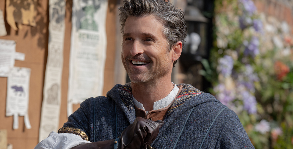 In a production still from Disenchanted, Robert (Disney Legend Patrick Dempsey) is outside, standing in front of a board that has announcements tacked to it; he’s looking off screen to the left and smiling wide. He’s wearing a blue tunic with a white shirt underneath, as well as brown gloves. The sun is shining.