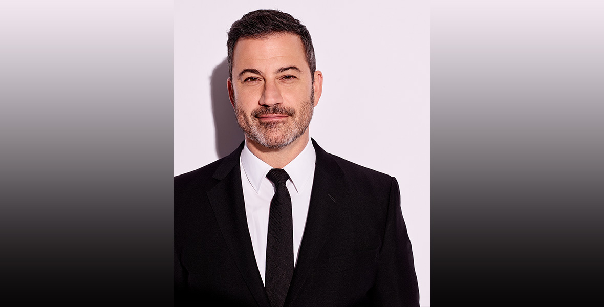 A headshot of Jimmy Kimmel; he’s wearing a black suit with a white shirt and black tie. He has dark hair and a salt and pepper beard, and is standing against a white background.