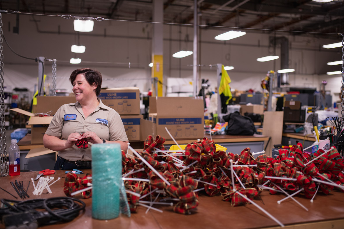 Cast member Carrie Henry stands behind a worktable in the Resort Enhancement warehouse in Anaheim, California. In front of her is a pile of the red-and-green bows she is making from ribbon.