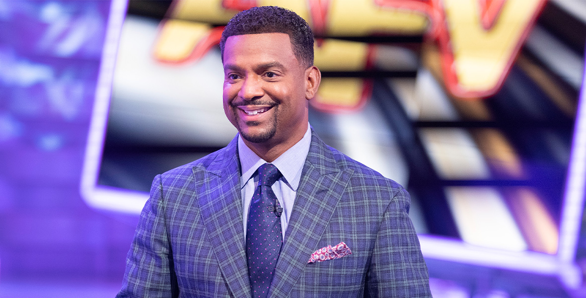 In an episode of America’s Funniest Home Videos, host Alfonso Ribeiro stands with his hands placed in front of him. He wears a blue and green plaid suit with a blue button-down shirt and a blue tie. Behind him is a large screen reading “AFV” and a stage adorned with bright blue lights.