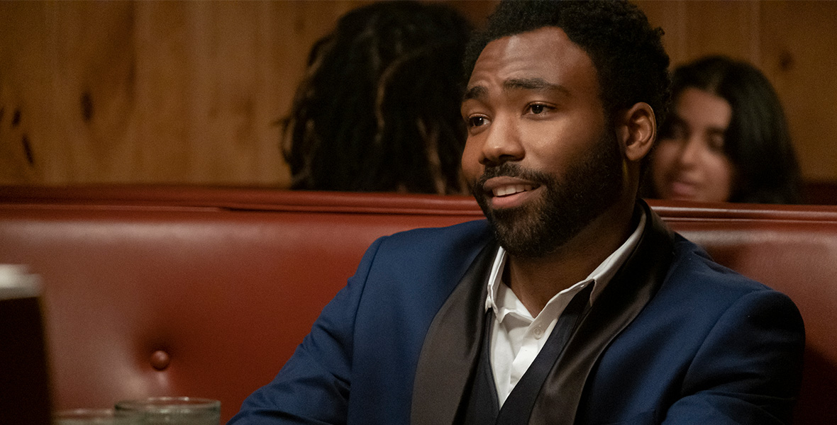 In a still from FX’s Atlanta, actor Donald Glover sits in a diner booth with orange leather seats. He wears a navy-blue tuxedo with black lapels and a white button-down shirt. The table in front of Glover holds a white plate and two clear water glasses.