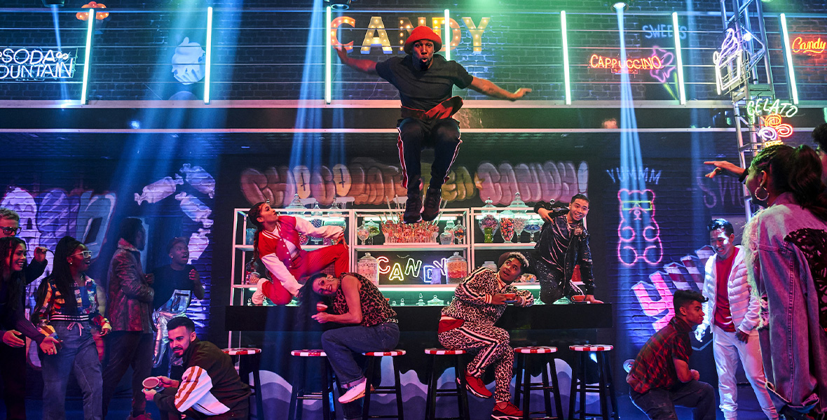 In a scene from Disney’s The Hip Hop Nutcracker, performer Stephen “Twitch” Boss jumps off the candy bar in the Land of Sweets. He wears a red hat, black T-shirt, and black pants with red stripes down the legs. Around him, the other dancers pose and look up and Boss who is in mid-air. Lights and decorations that read “CANDY” adorn the background and candy bar.