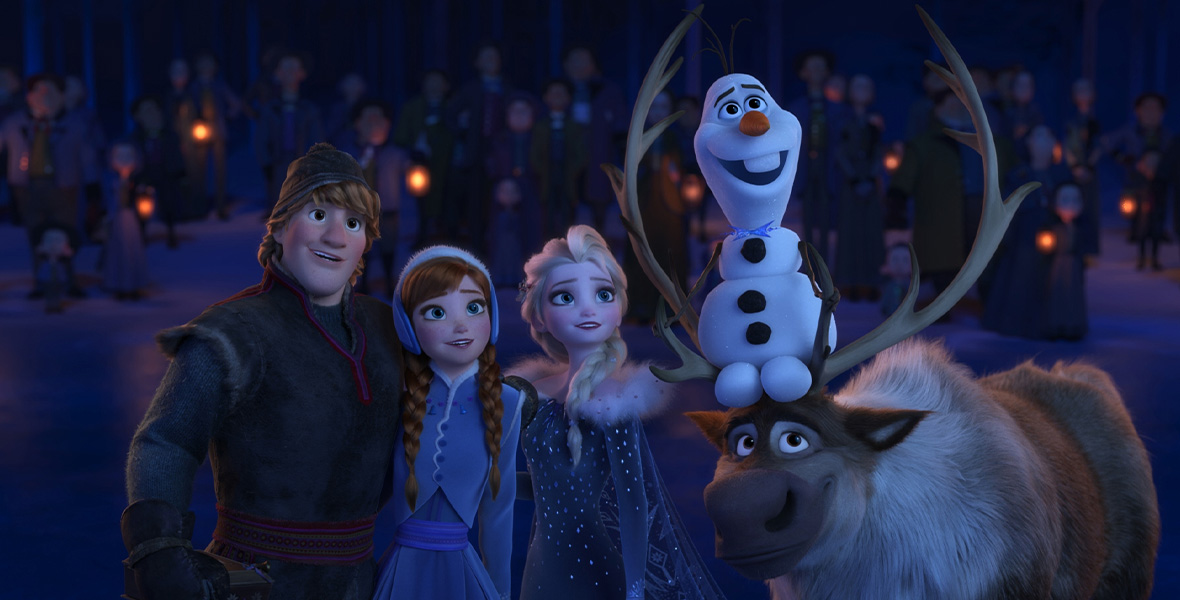 In a scene from Olaf’s Frozen Adventure, animated characters Kristoff, Anna, Elsa, Sven, and Olaf huddle together and look toward the sky. Kristoff wears a dark winter coat and matching pants with a beanie atop his head. Anna wears a periwinkle jacket and matching skirt with furry earmuffs. Elsa wears a dark blue gown covered in sparkles and a white fur neckline. Olaf, a snowman, sits atop Sven’s antlers. Behind them is a large group of people holding small lanterns.