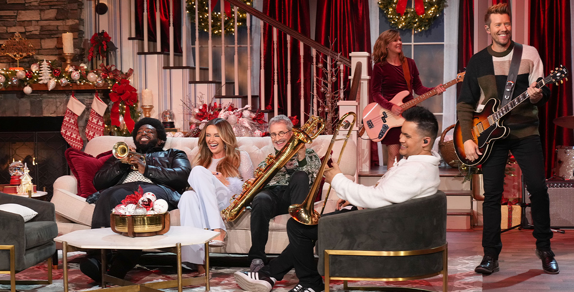 In a scene from CMA Country Christmas, host and country music performer Carly Pearce laughs as she sits center on a large white couch. Pearce wears a long-sleeve white top with feathers around the wrists, white pants, and white heels. On each side of Pearce sits a band member holding musical instruments—a trumpet and a saxophone. Another band member sits in an olive chair holding a trumpet while two guitarists stand to the side. Christmas stockings, garlands, candles, and decorations are on a staircase and fireplace behind them.
